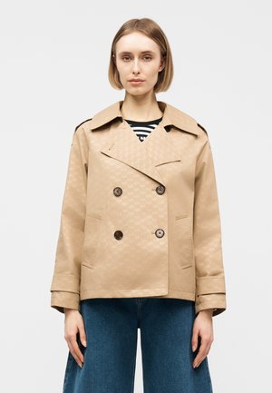 Γυναίκα με κοντά ξανθά μαλλιά φοράει μπεζ διπλομπρεδερό σακάκι με διακριτικό μοτίβο, ριγέ μπλούζα και μπλε τζιν παντελόνι φαρδιάς γραμμής, σε απλό φόντο.