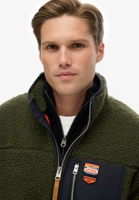 Fleecejack in olijfgroen met een marineblauwe stoffen inzet. Heeft een hoge kraag, een ritssluiting aan de voorkant en emblemen op de borst. Zachte textuur.