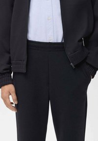 Persoon in zwarte elastische broek, zwarte ritsjack met elastische manchetten, en een witte blouse die is ingestopt.