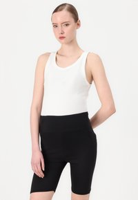 Witte geribbelde tanktop gecombineerd met hooggetailleerde zwarte fietsbroek, met een gladde textuur en een strakke pasvorm. Model poseert met een neutrale uitdrukking.