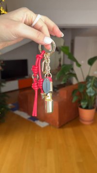 Μπρελόκ από κόκκινο δέρμα με χρυσά μεταλλικά στοιχεία, που διαθέτει ένα διακοσμητικό σφυρίχτρα και μια υφασμένη γοητεία. Το φόντο περιλαμβάνει φυτά εσωτερικού χώρου.