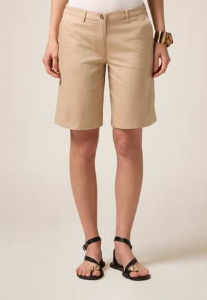 Bermuda beige con taglio dritto, chiusura frontale a bottoni e tasche, abbinate a sandali neri con pianta piatta e dettagli dorati.