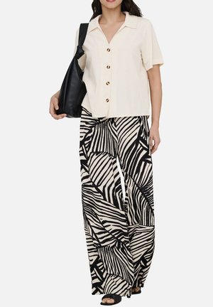 Femme portant une chemise beige à manches courtes boutonnée, un pantalon large à motif abstrait noir et blanc, des sandales noires et portant un sac à bandoulière noir.
