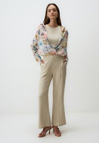 Blusa floreale trasparente con colori pastello, indossata sopra un top beige aderente. Pantaloni beige a gamba larga con cuciture frontali e sandali con tacco marroni.