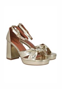 Sandalias de plataforma metálicas doradas con tacón grueso, con una correa frontal anudada y hebilla en el tobillo. Textura suave y diseño de punta redondeada.