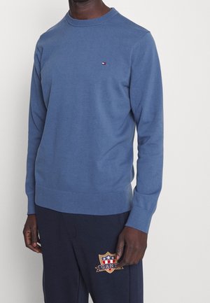 Man draagt een blauw trui met lange mouwen en een ronde hals, met een klein logo, en marineblauwe broek met een kleurrijk schildembleem.