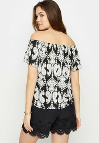 Top negro off-the-shoulder con estampado de paisley blanco, de tejido texturizado. Combinado con pantalones cortos negros que tienen bordes festoneados y recortes de flores.