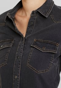 Musta farkk-shirtti, jossa on kaulus, erottuvan keltaisen ompeleet, kaksi rintataskua ja nepparit. Pintarakennedetali näkyvissä.