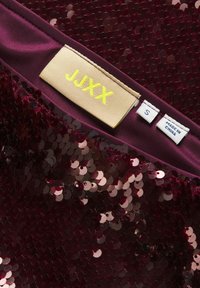 Haut en tissu bordeaux orné de sequins superposés, avec une étiquette jaune portant la mention "JJXX", la taille "S" et un label "Fabriqué en Chine".