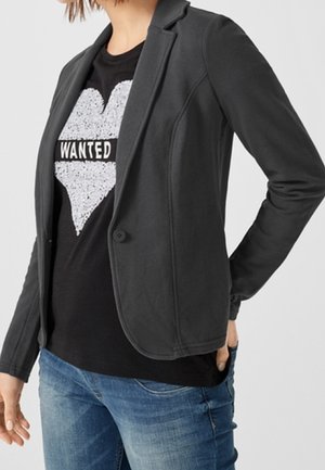 Person wearing a black blazer over a black T-shirt with a white heart and the word "WANTED," paired with blue jeans.