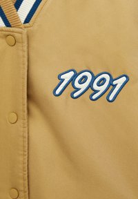 Kurtka w kolorze tan z zatrzaskami, z wyszywanym białym i niebieskim napisem "1991" na klatce piersiowej oraz w paskowanym kołnierzu w kolorze niebieskim i białym.