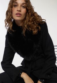 Cappotto nero con colletto in pelliccia sintetica, caratterizzato da un design strutturato, maniche lunghe e vita legata. Gli accessori includono piccoli bottoni sui polsini.