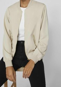 Personne portant une veste beige à fermeture éclair par-dessus une chemise blanche avec un pantalon noir, assise sur un tabouret en bois devant un fond neutre.