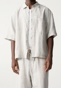 Homme portant une chemise à manches courtes beige clair ample boutonnée et un pantalon assorti, avec un débardeur blanc côtelé en dessous, debout devant un fond blanc.