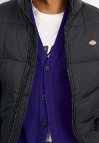 Svart pufferjacka med dragkedja, bärs över en lila cardigan med knappar. En vit T-shirt syns under.