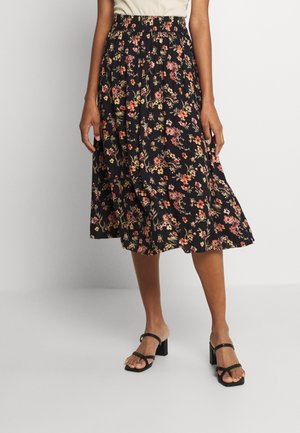 Persoon draagt een zwarte midi-rok met bloemenmotief in roze en geel, een beige top en zwarte sandalen met hak, tegen een effen achtergrond.
