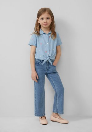 Chemise bleu clair à manches courtes avec broderies colorées, nouée à la taille, associée à un jean en denim taille haute et des ballerines rose pâle.