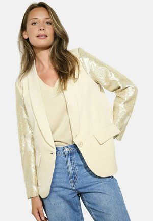 Blazer crema con maniche a motivo dorato, chiusura con un bottone e due tasche frontali. Abbinato a una maglietta oro chiaro e jeans blu.
