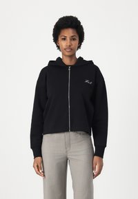 Schwarzer, kurzer Hoodie mit einem Frontreißverschluss und Kapuze mit Kordelzug. Verfügt über gestickten Text auf der Brust und graue, strukturierte, weite Hosen.