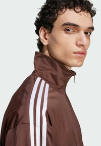 Veste brune zippée avec col haut, rayures verticales blanches sur les épaules, texture lisse, design athlétique, avec une coupe élégante.