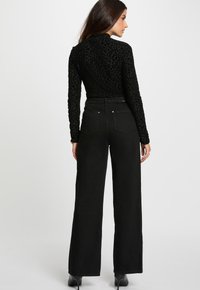 Donna che indossa pantaloni larghi a vita alta neri, una maglietta a maniche lunghe con stampa leopardata nera e stivali con tacco neri, in posizione di profilo.