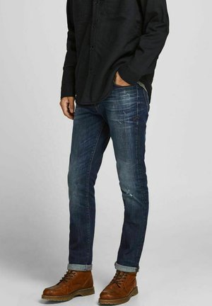 Mann trägt hochgekrempelte blaue Jeans, braune Leder-Schnürstiefel und ein langärmeliges schwarzes Hemd, eine Hand in der Tasche.