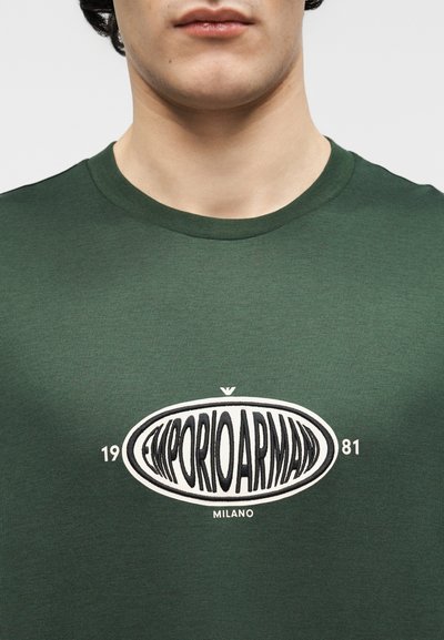 Κοντινό πλάνο ενός ατόμου που φοράει μπλουζάκι σκούρου πράσινου χρώματος με λευκό οβάλ λογότυπο που γράφει "EMPORIO ARMANI 1981 MILANO" στο στήθος.