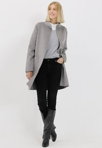 Grijze wollen jas met een hoge kraag en twee knopen, gecombineerd met een grijze trui en zwarte skinny jeans, aangevuld met kniehoge zwarte laarzen.