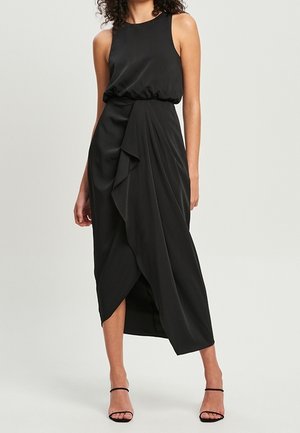 Vrouw draagt een mouwloze zwarte midi-jurk met een geplooide taille en gedrapeerde asymmetrische rok, gecombineerd met zwarte sandalen met hoge hakken en bandjes.