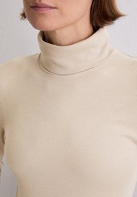 Beige gerippter Rollkragenpullover mit tailliertem Schnitt und weicher Textur, der einen hohen Kragen besitzt, der über den Hals reicht.