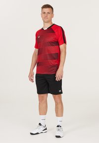 Lyhythihainen urheilupaita, jossa on punamustaraitoja ja V-kaula-aukko, yhdistetty mustiin shortsien kanssa. Mallilla on valkoiset urheilukengät ja sukat.