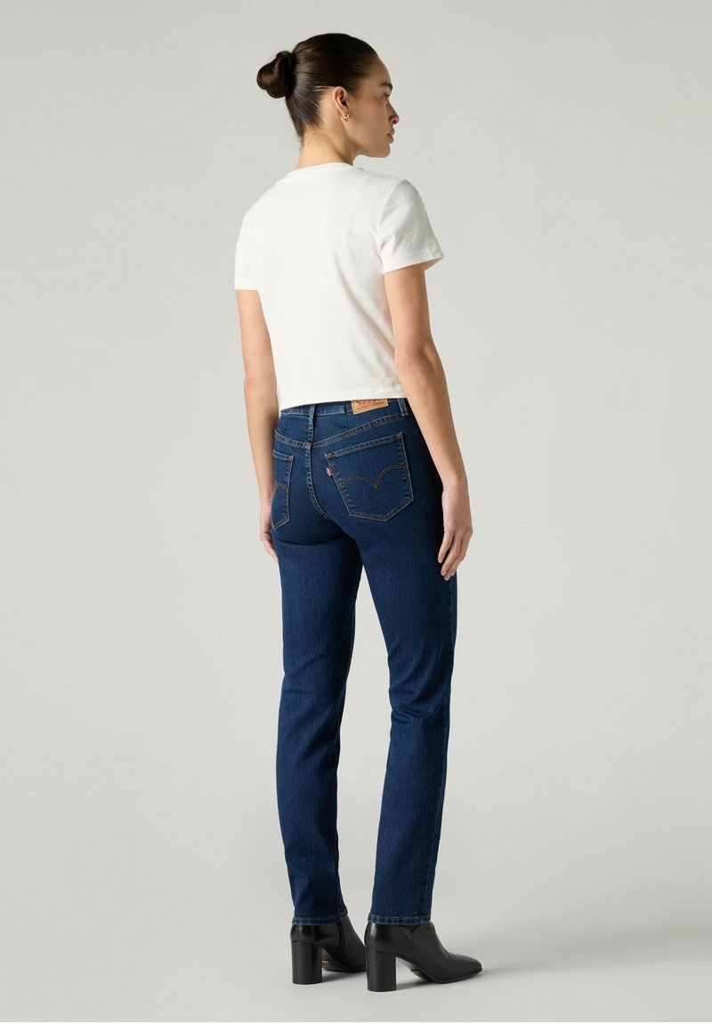 Femme avec un chignon, portant une chemise blanche à manches courtes, un jean bleu foncé et des bottines noires à talons, debout de profil.