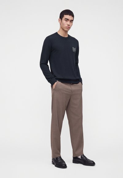 Homme portant une chemise noire à manches longues, un pantalon marron et des mocassins noirs, debout les mains dans les poches sur fond blanc.