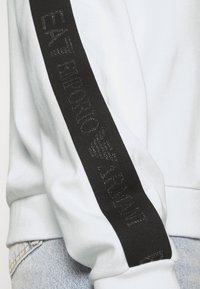 Manica di felpa bianca con striscia nera che presenta un testo sottile "EA7 Emporio Armani", indossata con jeans di denim azzurri chiari.