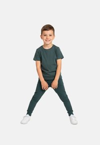 Ciemnozielona koszulka i dopasowane spodnie dresowe wykonane z miękkiego materiału. Dziecko stoi z nogami rozstawionymi, w białych tenisówkach, uśmiechając się. Prosty design.