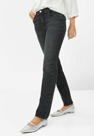 Femme portant un jean noir slim, une blouse blanche et des ballerines argentées à bout pointu avec des brides à la cheville, debout avec une main dans la poche.