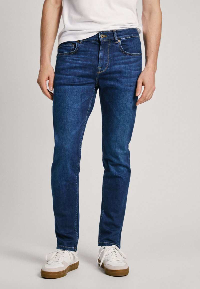 Jeans de mezclilla azul oscuro con un corte slim, que presentan un diseño clásico de cinco bolsillos, costuras visibles y un cierre frontal de botón.