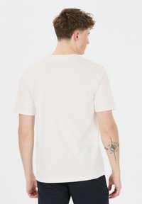 Bílý bavlněný tričko s krátkým rukávem, jednoduchý design a kulatým výstřihem, které nosí osoba ukazující tetování na pravém rukávu.