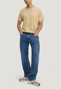 Krátké béžové bavlněné tričko s kulatým výstřihem, doplněné volnými modrými džínami a světle hnědými sportovními botami.