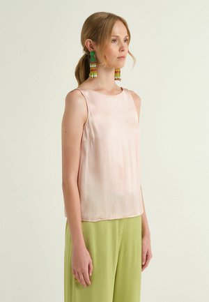 Femme aux cheveux blonds attachés en queue de cheval, portant un haut sans manches rose pâle, un pantalon vert clair, et de longues boucles d'oreilles perlées vertes et roses, tournée vers la droite.