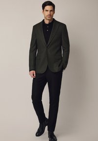 Ciemnozielona marynarka z klapami o zakładkach i dwoma przednimi kieszeniami, zestawiona z czarną koszulką polo i czarnymi eleganckimi spodniami, dopełniona czarnymi butami.