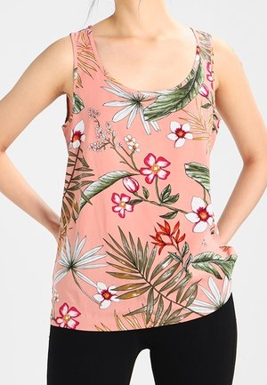 Vrouw draagt een mouwloze roze top met groene en witte tropische bladeren- en bloemenprint, gecombineerd met een zwarte broek.