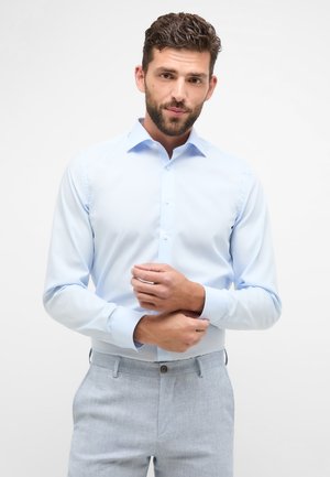 Chemise de couleur bleu clair avec des manches longues, fermée par des boutons à l'avant et aux poignets. Le tissu semble lisse avec une coupe ajustée. Style léger et formel.