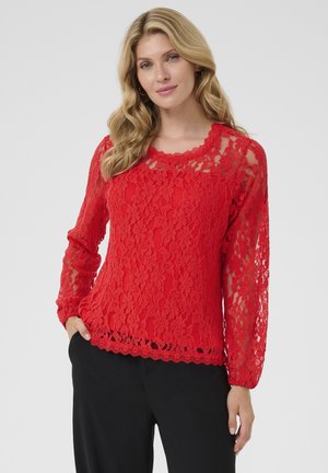 Red lace top featuring long sleeves, scalloped neckline, and a textured pattern with a straight hem. Paired with black trousers.