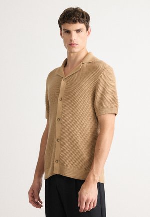 Young man wearing beige short-sleeve knit button-up shirt with textured pattern and black pants, standing against plain white background.