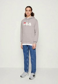 Sweatshirt cinza com o logótipo FILA em branco e vermelho, bolso canguru, sobre uma camisa branca, emparelhado com jeans azuis e ténis cinzas.