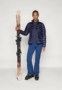 Chaqueta acolchada azul marino con diseño de acolchado vertical, combinada con pantalón de esquí azul y botas de esquí negras, sosteniendo un esquí con fijaciones.