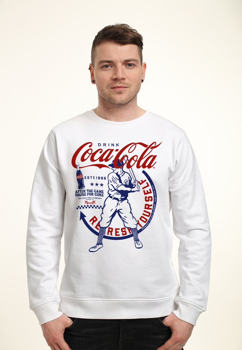 Weißer Hoodie mit einem Vintage Coca-Cola Grafik. Das Design zeigt einen Baseballspieler in Rot und Blau mit Text und Akzenten.
