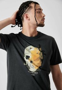 T-shirt en coton noir avec un graphique de skull vieilli en jaune et blanc, avec du texte en dessous. Encolure ronde et manches courtes.