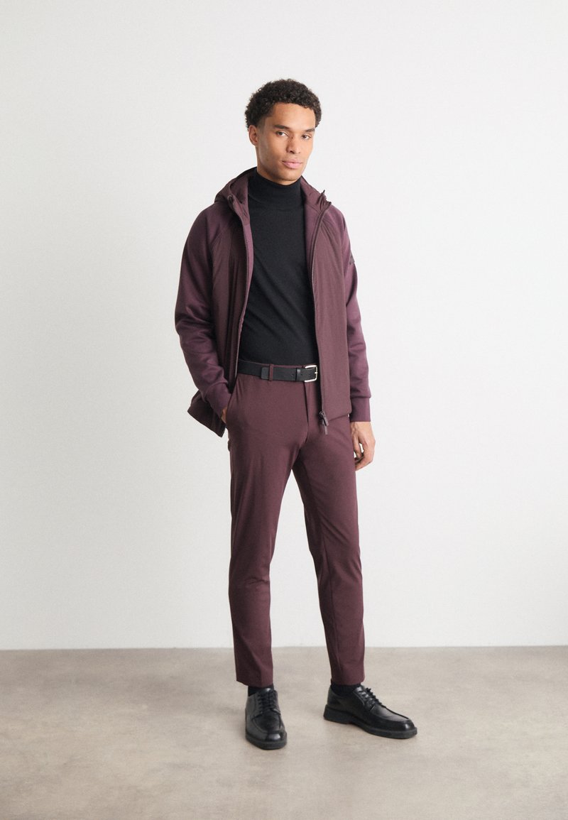 Burgunderrote Jacke über einem schwarzen Rollkragenpullover, kombiniert mit burgunderroten, taillierten Hosen und schwarzen Schuhen. Glatte Textur, figurbetontes Design, minimaler Einsatz von Hardware.